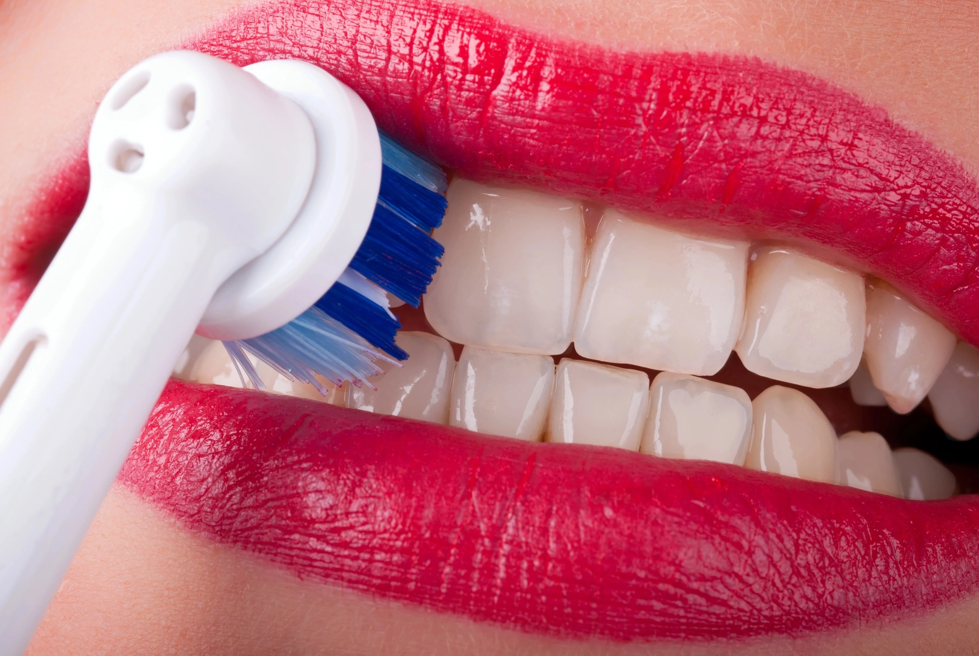 Close-up of a mouth with bright red lipstick, demonstrating tooth brushing with an electric toothbrush on sparkling white teeth from a dentist on hiatus rd