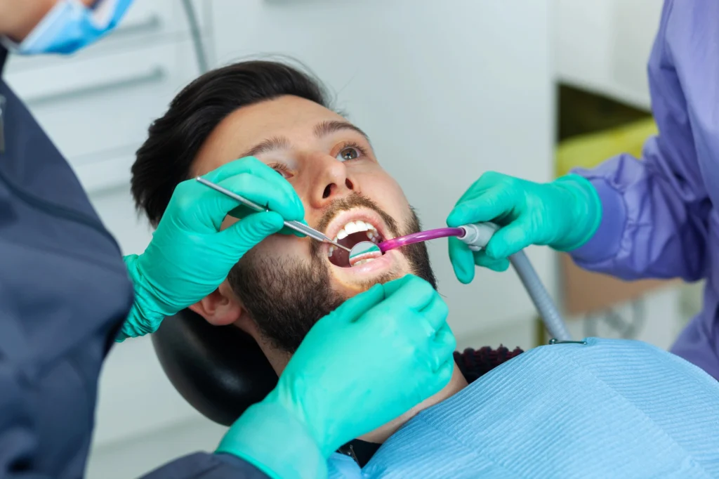 A dentist on hiatus rd in green gloves uses a dental tool on a patient reclining in a chair, wearing a blue bib in a clinical setting.